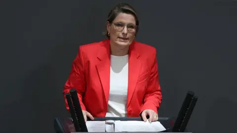 Sean Gallup/Getty Images A woman in a red jacket and white T-shirt talks in front of an audience of MPs in Berlin