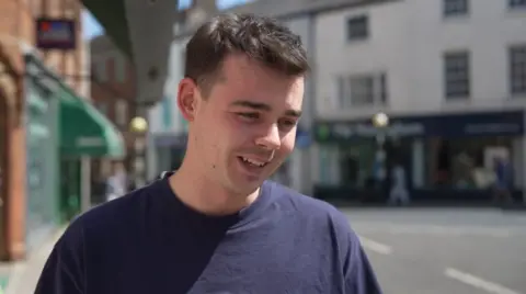 A young man standing in the street. He has dark hair and is smiling. There is a row of shops in the background