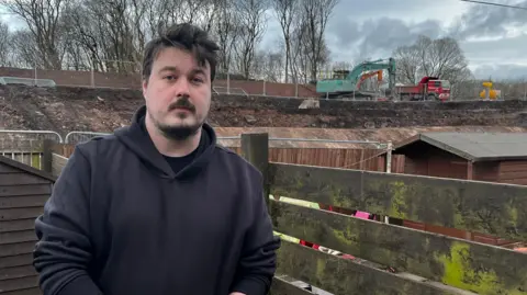 A man with dark hair and facial hair, wearing a black hoodie standing in front of a garden shed and fence.