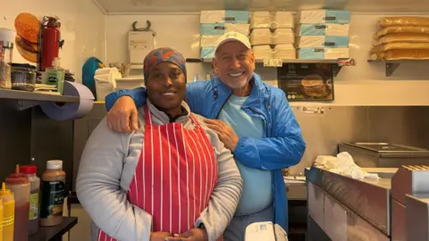 Violet John and James Nas, the owners of the Hot Sausage Company, standing in their van. They are both smiling into the camera. Violet is wearing a red and white apron and James is wearing a blue jacket.