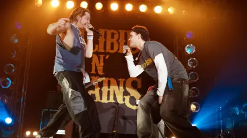Two rappers in late 90s/early noughties clothing performing on stage with a backdrop of bright lights and a large sign that reads "Silibil N' Brains"