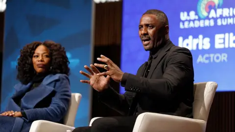 Getty Images Idris Elba (right) gestures as he speaks onstage while sitting next to Yvonne Orji (left), with a blue projector screen in the background that says 'US Africa Leaders Summit' and 'Idris Elba, actor' on it