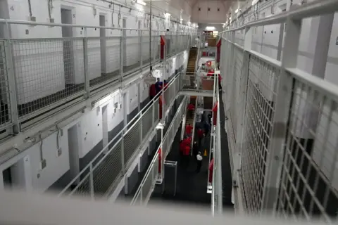 a view from top to bottom of the shite metal fences of the landings, from third floor to ground, with prisoners in red outfits down below.