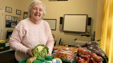 An older lady with grey hair wearing a pink knitted jumper. She is smiling, holding a cauliflower. She's stood in a room with lots of food on a table including tomatoes and soup. There are certificates and photographs on the walls behind her. 