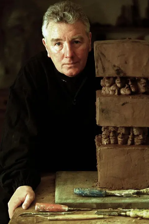 Getty Images Jimmy Boyle, dressed in black clothing, stands next to a three-tiered clay sculpture. It is placed on a table, with tools nearby.