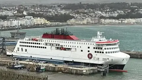 The Manxman ferry moored in Douglas Harbour. It is a large vessel painted in the Isle of Man Steam Packet Company's colours of white, red and black, with the firm's logo featuring the three Legs of Mann in a circle on the side. Douglas seafront can be seen in the background.