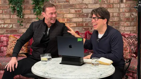 Emily's Duncan Ferguson and Michael McIntyre, who are both laughing in the picture, are sitting at a table in Emily's restaurant. There are both looking at a Mac book and there is a three glasses filled with water on the table and a notebook.
