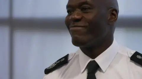 The man in the picture is smiling at something off-camera. He is wearing a white shirt and black tie with black epaulettes.