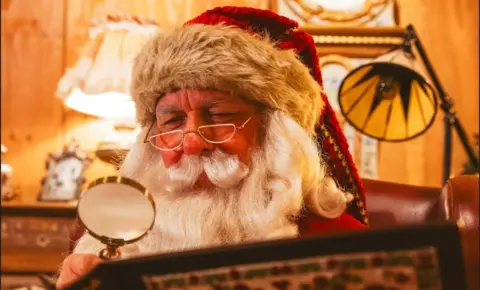 Lapland UK Santa who has a traditional red and white hat, white beard with half rimmed glasses is holding a magnifying glass over his Christmas list.