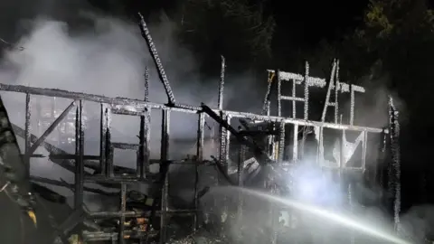 Photo shows the burnt out shell of a derelict one storey building. All that remains is a charred black timber frame.