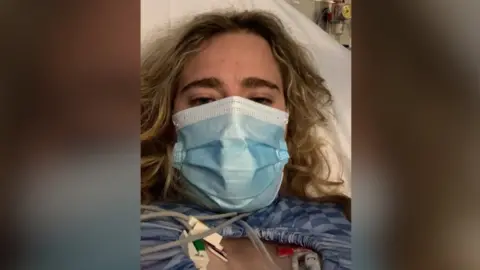 Zoe Hamer Picture of a blonde woman wearing a surgical mask and lying in a hospital bed.