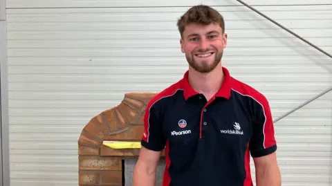 Joe Shingler, wearing a WorldSkills black and red polo shirt. He has short ginger hair and beard and is smiling at the camera against a white backdrop, with an example of his brickwork behind him. 