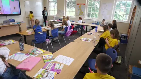 Children wearing T-shirts sit in a classroom in front of desks. A teacher stands at the front and is instructing them. A child writes on a whiteboard and a monitor is displaying an insect. Textbooks can be seen on some of the desks. 