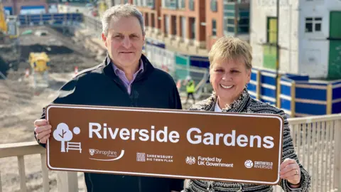 Deputy council leader Ian Nellins and council leader Lezley Picton stand next to the site, which is currently being cleared. They are holding a sign showing the park's new name.