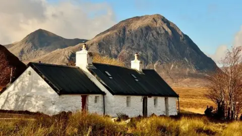 There is a white house with a dark roof. It is surrounded by yellow long grass. In the background is a mountain.