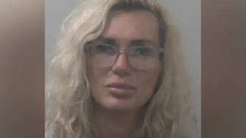 Kasha Sanderson has wavy blonde hair and is wearing glasses and makeup in a police mugshot.