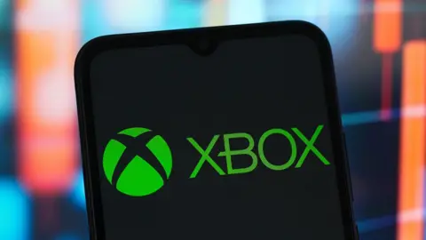 Xbox logo displayed on a smartphone screen, in front of a backdrop of a blurred financial chart with red candlestick indicators.