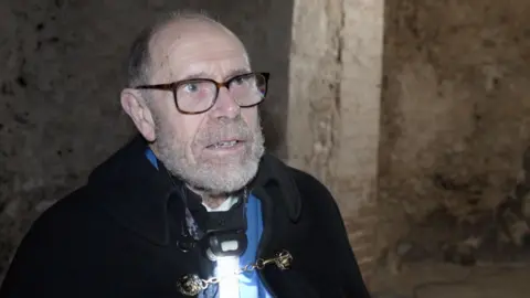 Shaun Whitmore/BBC Tony Mills, wearing a black cloak and brown glasses. He is looking off to the side of the camera.