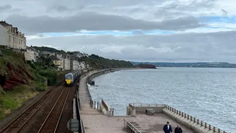 Sea wall in Dawlish. There are train tracks and a train on the left, and a pathway on the right. To the right of the trainline are houses and a cliff face.