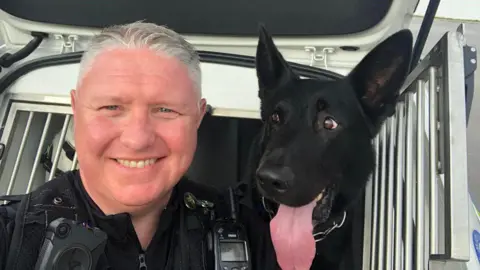 A police officer smiling at the camera with a black German shepherd, who is sticking his tongue out