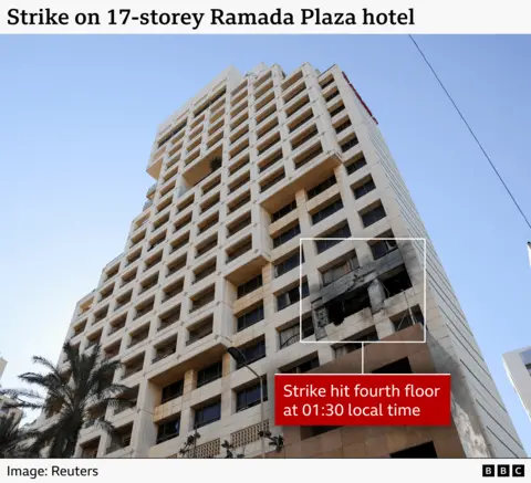 A tall, beige, concrete high‑rise hotel building stands against a clear blue sky. The structure has 17 storeys, with a grid-like façade made up of repeating square window openings framed by thick vertical and horizontal beams. Near the lower-middle section of the building, roughly at the fourth floor, a section of the façade is visibly damaged: the windows are blown out, and the surrounding concrete is darkened and charred. A white rectangular outline highlights this damaged area. At the bottom right of the image, a red label reads “Strike hit fourth floor at 01:30 local time.” 