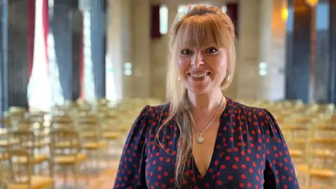 Delyth is wearing a navy dress with red spots and is smiling at the camera. She is stood in a large function room with rows of chairs in the background. She has a blonde fringe, with her long hair tied back.