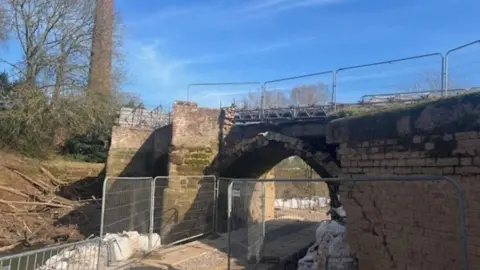 Worcestershire County Council Closed bridge, with barriers on top of it and also near it close to the bottom of the bridge by the river.