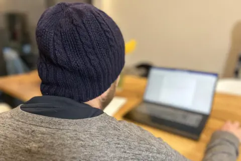 A man wearing a blue beanie hat and a grey sweater is pictured from behind. He is looking at a laptop