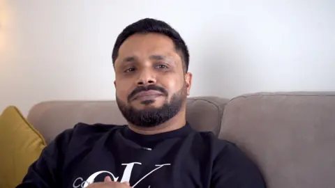 Kamrul Husan Sumon wearing a black Calvin Klein jumper sitting on a light brown sofa in his lounge. He is looking directly at the camera.