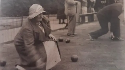 Contributed A black and white image of a woman bowling on a green. She is crouched as she bowls. She wears a bowler hat and a coat and dress. 