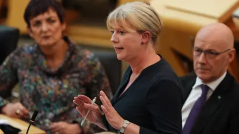 Shona Robison, a woman with blonde hair, speaks in the Scottish Parliament chamber. She is wearing a black dress and has her hands out in front of her chest. 