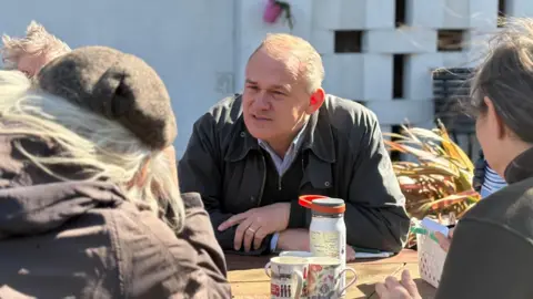 LDRS Ed Davey is sitting at a table as he talks to a man and a woman. He has short grey hair and is wearing a khaki jacket over a black jumper. There are two mugs and a water bottle on the table. 