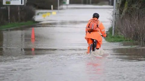 A man wearing orange wet weather gear riding his cycle through surface water on a road.