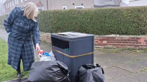 Joan McTigue is wearing a long blue coat and looking at a black bin surrounded with black bin bags full of rubbish. 