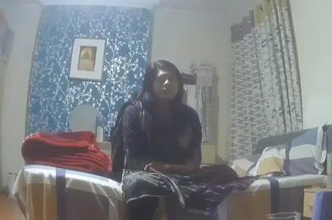 Undercover footage of an Asian woman sitting on a bed. The chimney breast behind her has been turned into a feature wall with bright blue and silver wallpaper, while the bed cover is a large check pattern in yellow, blue and grey.