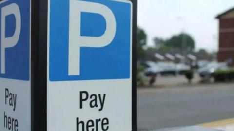 A parking meter sign with a while capital P inside a blue box, and black text on a white background below saying 'Pay here'. A car park is in soft focus in the background