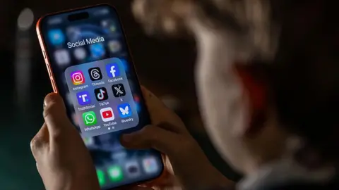Close-up of a person holding a smartphone displaying a folder of social media apps, including Instagram, Facebook, TikTok and WhatsApp.