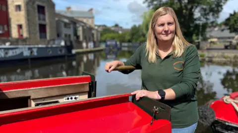 Olivia Richwald/BBC A woman with long blonde hair stands at the back of a red narrow boat. She is holding onto a tiller and you can see the canal and another narrow boat in the distance. It is sunny.