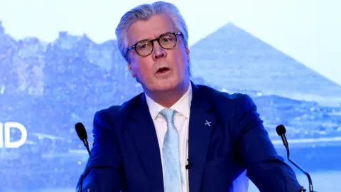 Malcolm Offord, who has grey hair and dark-rimmed glasses, speaks at a podium in front of a blue Reform-branded sign 