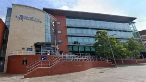 The outside of the university building. It has a glass frontage with trees in front of it on the right and steps leading up to the entrance on the left.
