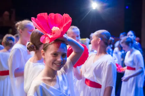 Jane Barlow/PA Wire A young Ukrainian girl in a white dress smiles and holds a big red flower over her head while on stage. Similarly-dressed children stand in a group behind her under the theatre lights.
