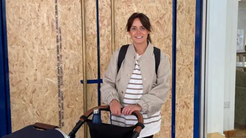 Rebecca Turner stands in front of doors with boards on with a pram