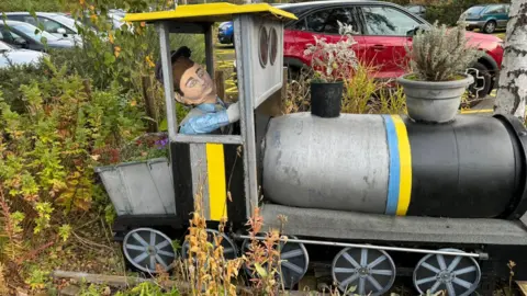 Een grijs, zwart en geel model van een trein met een opgezette figuur in de cabine en plantenpotten als trechter