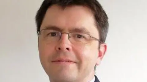 Headshot of David Hill. He has short brown hair and is wearing glasses, looking into the camera.