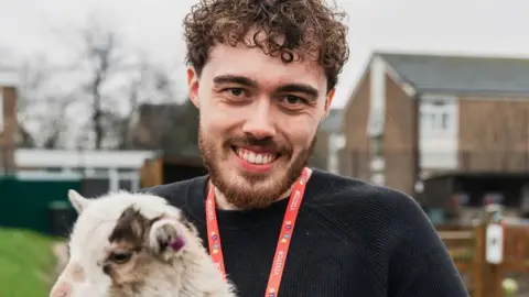 BILTT/Grange Academy Toby, holding a goat in his arms, with a red lanyard round his neck, wearing a black jumper, with a beard and short curly hair. School buildings, that are blurred are behind him. 