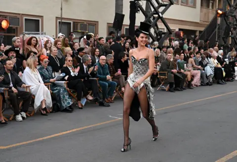 Getty Images Kendall Jenner wearing a crystal encrusted bodysuit and top hat. She is smiling and walking along the outdoor catwalk