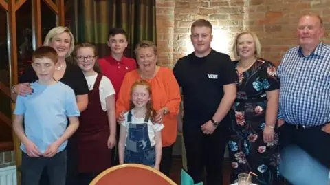 McGrath Family June McGrath posing for a photo surrounded by family members of various ages. The pensioner is smiling and has her hands resting on the shoulders of a young girl with long, ginger hair. Ms McGrath is wearing a bright orange top. 