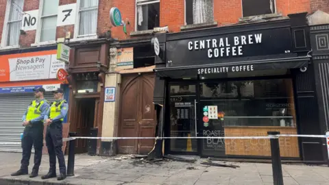 Two gardai officers dressed in uniform standing on the street outside a shop and coffee shop where the damage caused by the fire to the prayer hall can be seen. the area is cordoned off.