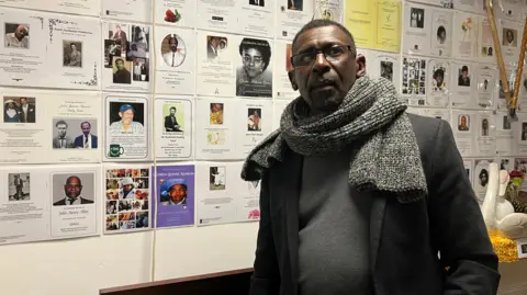 Jon Wright/BBC Max Thomas with a coat and scarf, standing in front of a display board with lots of photos of people