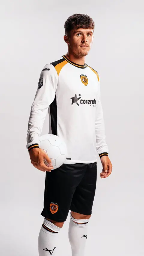 Hull City A man wearing a Hull City away kit stares into the camera. He is holding a white football in one hand. The kit features a white shirt with black and amber trims on the front of the shoulders, collars and seams. Black shorts and white socks complete the outfit.